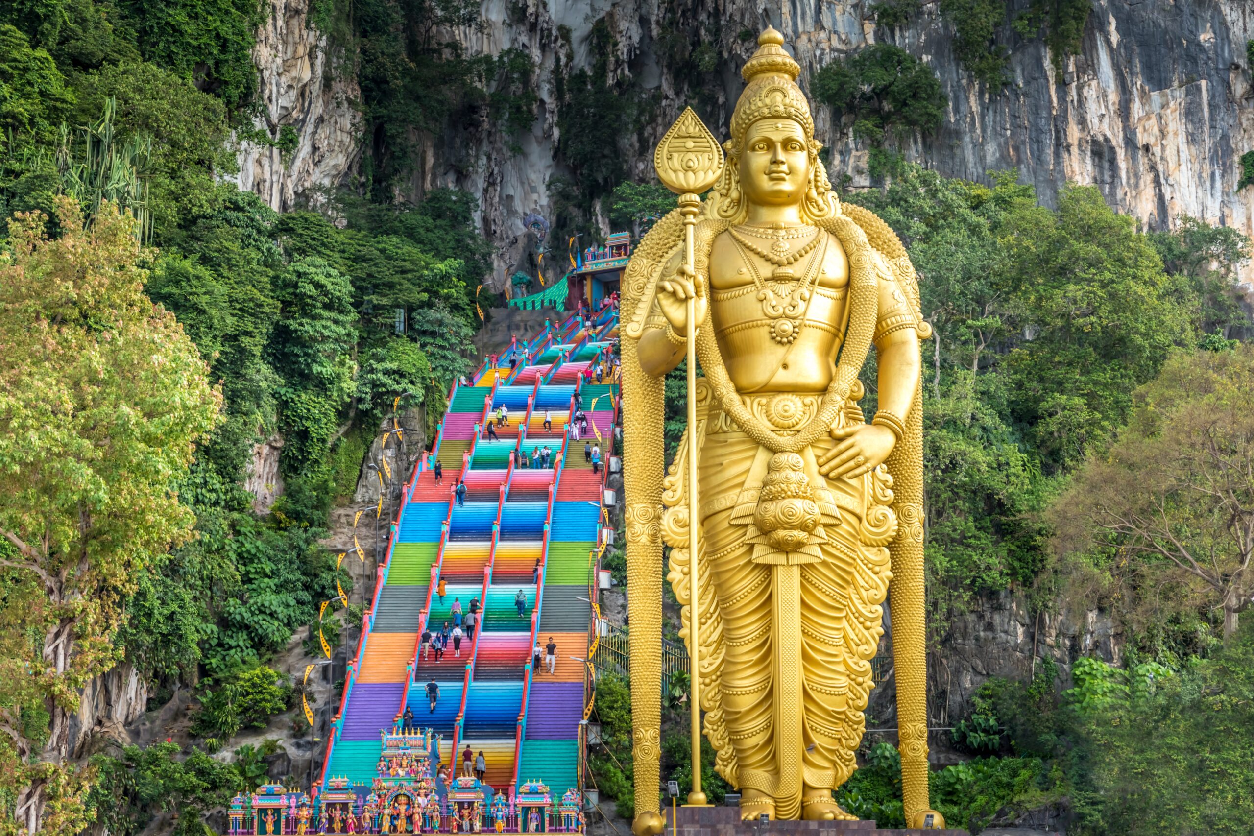 Large golden statue of a Hindu deity beside a bright, multicolored staircase at Batu Caves with a rocky cliff in the background.