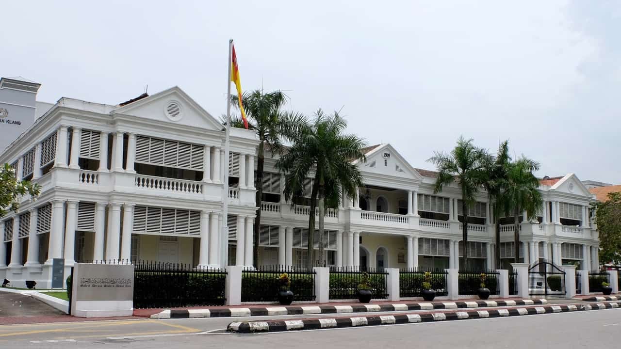 White colonial-style government building with two stories, tall columns, balconies, and a flagpole with a red-yellow flag, fenced by a black gate and palm trees nearby.